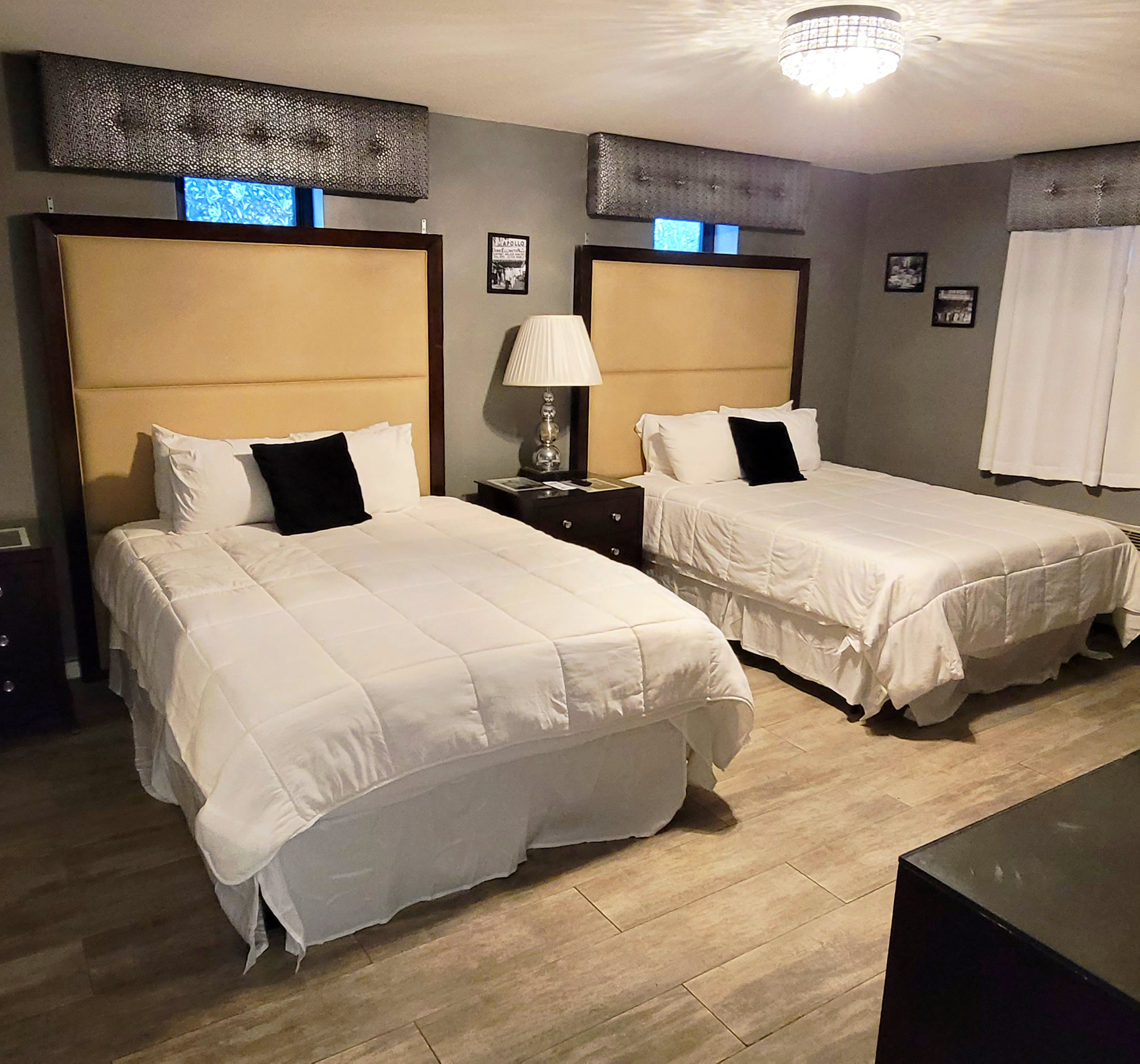 2 Queen size beds with white sheets 2 white pillows and one black pillow with a tan tufed headboard in a room with tan carpet and grey walls.