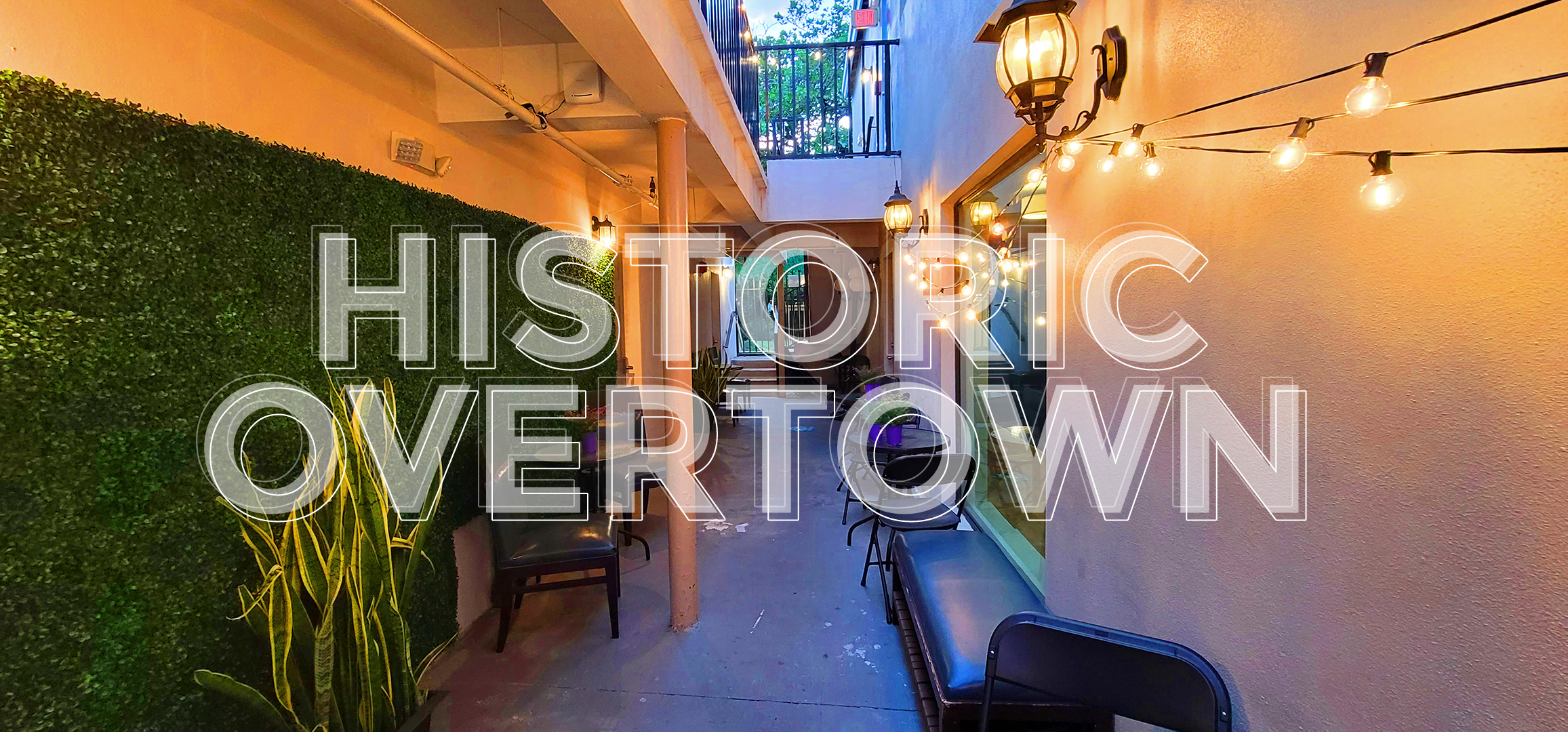 Image of exterior walkway with artificial greenery on left wall with string light on the right wall with the words HISTORIC OVERTOWN in the center.